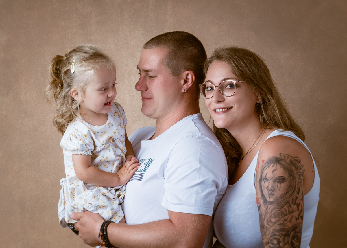 Junge Familie vor warmem Hintergrund: Vater hält die kleine Tochter im Arm, während die Mutter daneben lächelnd steht.